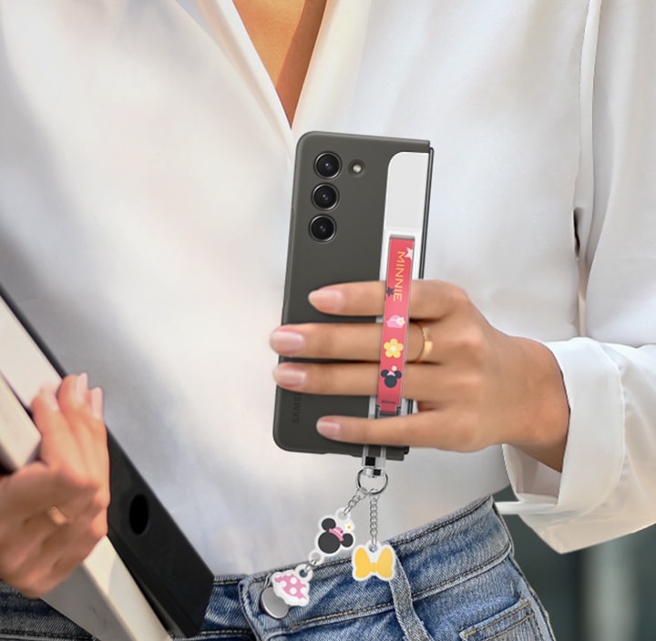 Une jeune femme en tenue semi-formelle marche à l'extérieur, tenant un smartphone Galaxy Z Fold5 avec une coque avec module de maintien interchangeable, ses doigts glissés sous une lanière de couleur rouge. Des accessoires colorés sont attachés à la partie inférieure de la coque. 