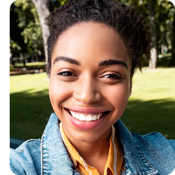 Selfie of a young smiling woman looking into camera lenses, with a little bit of green grass and tree shown in the background