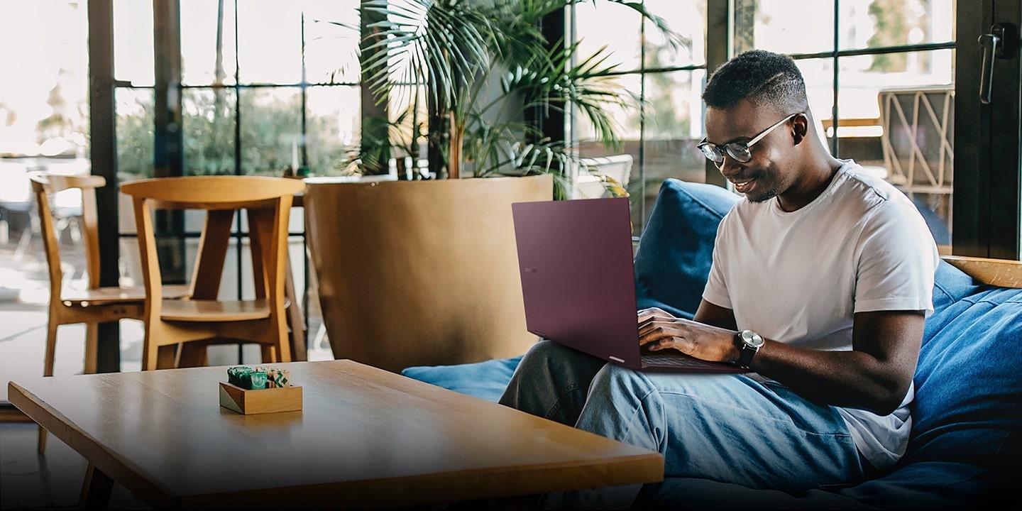 Aan de rechterkant zat een man op een grote blauwe pouf in een zonnige kamer met een grote plant en houten meubels omgeven door glazen wanden. De man gebruikt een antraciet Galaxy Book2 Pro 360 op zijn knieën, glimlachend