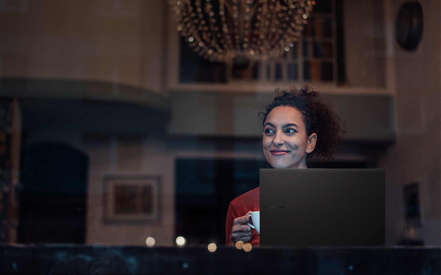 Uma mulher está sentada em uma sala de estar luxuosamente decorada com um lustre e obras de arte. Enquanto ela segura uma xícara na mão direita, atrás do Galaxy Book2 Pro, ela olha para a esquerda sorrindo