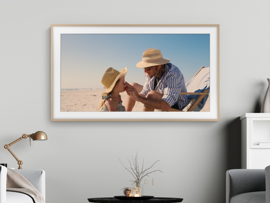 Een vader raakt het gezicht van zijn dochter aan op het strand. Het moment wordt vastgelegd op het frame, dat aan de muur van een kamer wordt opgehangen