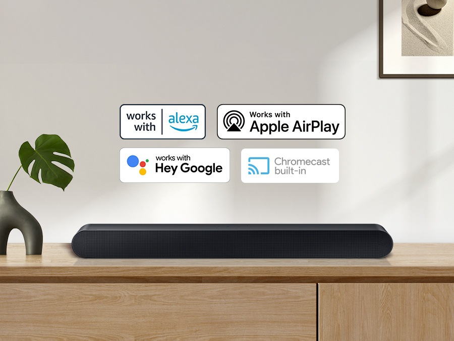 Le logo Alexa, le logo AirPlay d'Apple, le logo Hey Google et le logo Chromecast Built-in peuvent être vus avec la barre de son Samsung S60B qui est posée sur le meuble du salon.