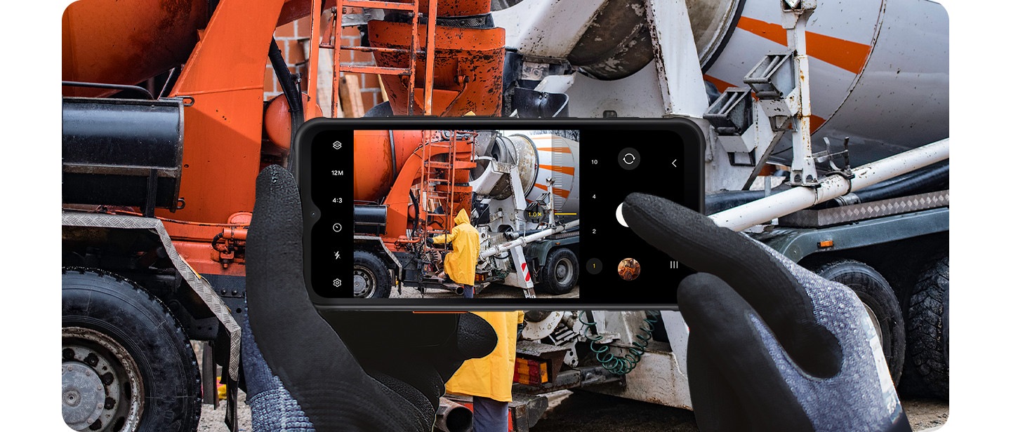 Deux mains portant des gants de protection tiennent le Galaxy Xcover7 tout en utilisant l'application appareil photo pour capturer une scène sur un chantier de construction.