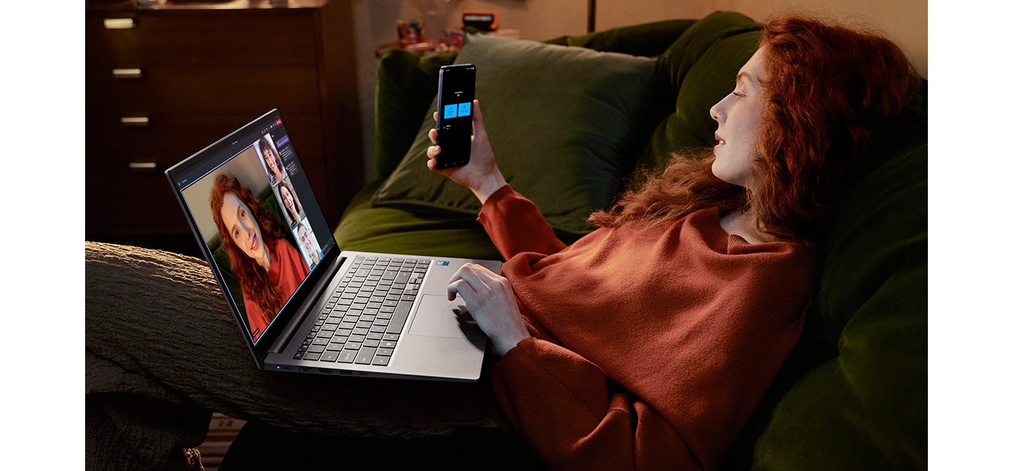 Une jeune femme assise sur un canapé à la maison utilise le Galaxy S24 Plus comme caméra connectée avec le Galaxy Book4 à Gray pour participer à un appel vidéo de groupe Microsoft Teams avec trois autres personnes affichées à l'écran.