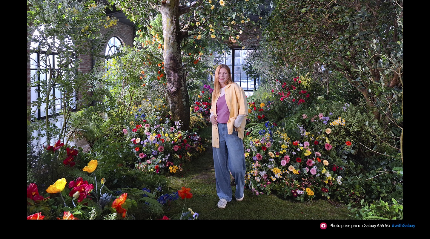 A photo taken with 50 Megapixels of high-resolution of a person standing in a lush indoor garden full of colorful flowers and greenery. Text reads Captured by Galaxy A55 5G #withGalaxy.