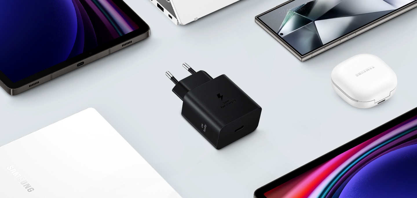 Une vue de dessus de divers appareils Samsung Galaxy est présentée avec un adaptateur d'alimentation noir placé au centre. À gauche, un Galaxy Book3, à droite un Galaxy Buds2 Pro en blanc, un Galaxy Tab S9 et un Galaxy S24 Ultra sont également montrés.