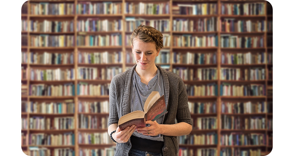 1. A woman reading a book standing in front of a bookcase full of books. The bookcase in the background in blurred out.