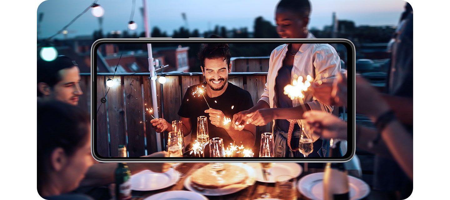 Galaxy A72 in landscape mode. Onscreen is a scene of people at a party and it expands outside of the display. The image inside is brighter and more detailed, demonstrating OIS.