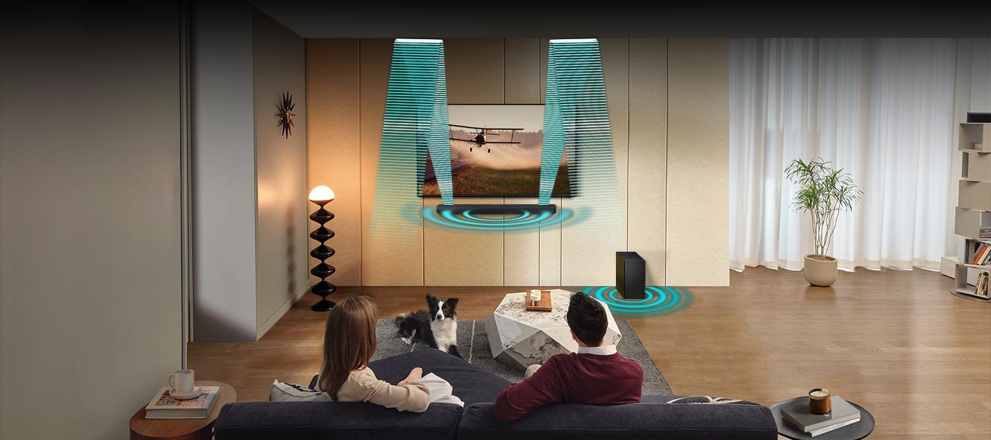 In a living room, a couple sits on a sofa and watches TV. The TV is installed onto the wall, with a Soundbar and subwoofer underneath. The Soundbar gives off angled sound waves that travel up, hit the ceiling, and bounce down to the back of the room. Then round sound waves emanate from both the Soundbar and subwoofer.