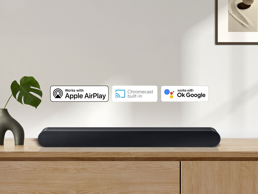 Apple AirPlay logo, Chromecast Built-in logo, and Ok Google logo can be seen along with Samsung S60B soundbar which is sitting on living room cabinet.