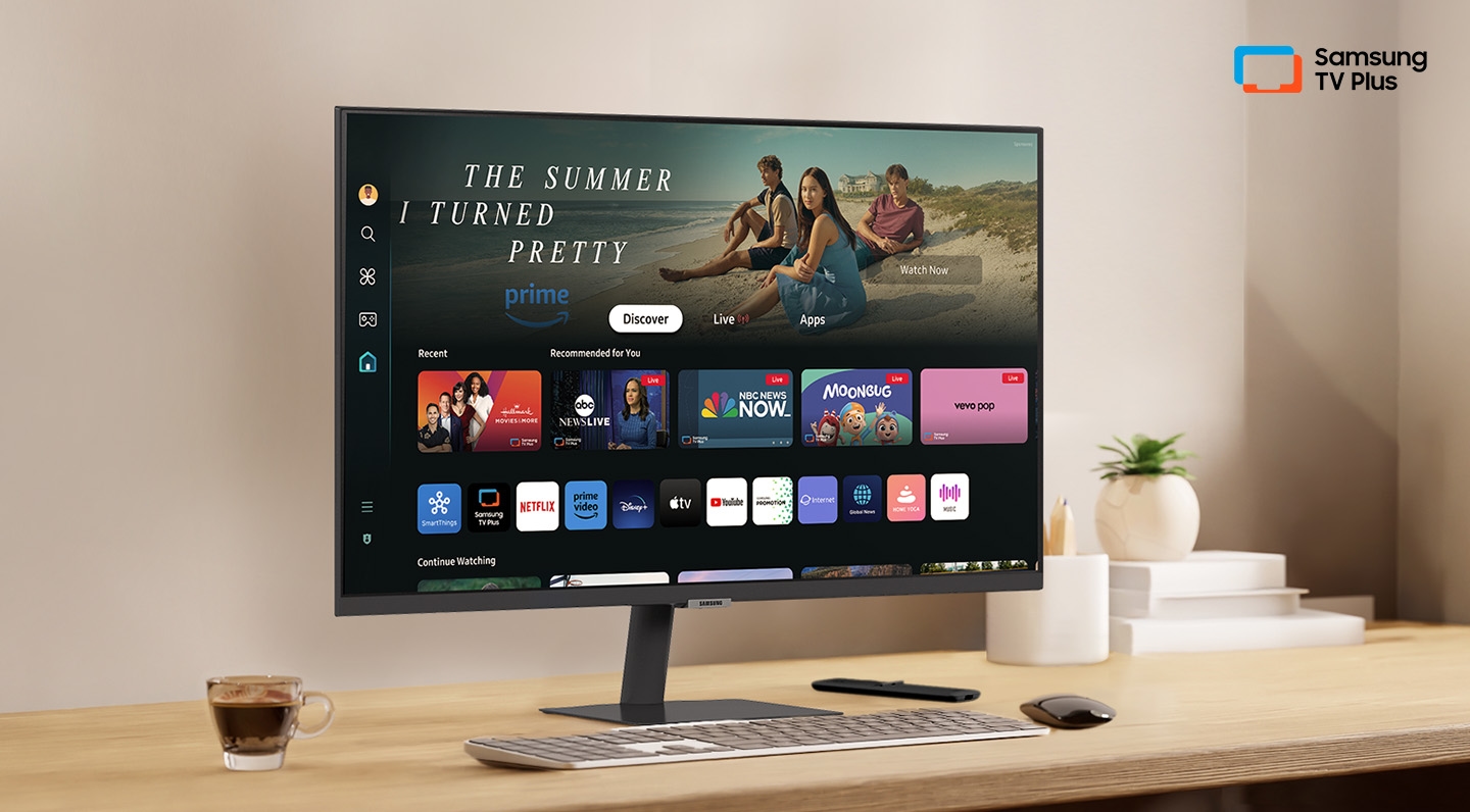 There is a monitor on a desk and its home UI is shown with OTT applications. On the upper right, there's a logo of Samsung TV Plus.