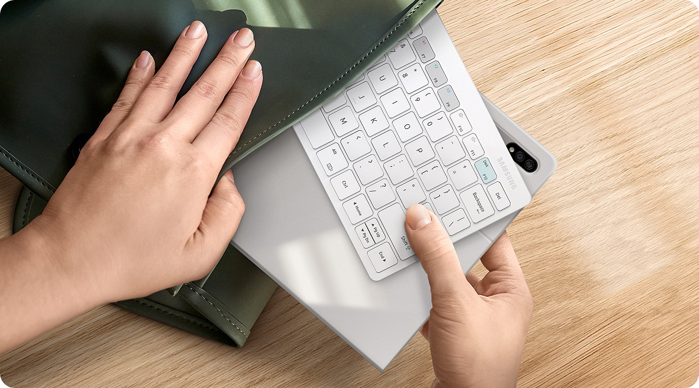 There is a green bag on a desk and inside of the bag there is a white-colored Samsung Smart Keyboard Trio 500 and a tablet that are halfway inside. One hand is grabbing the keyboard and the tablet, and the other hand is grabbing the bag to take out the devices or put them back in.