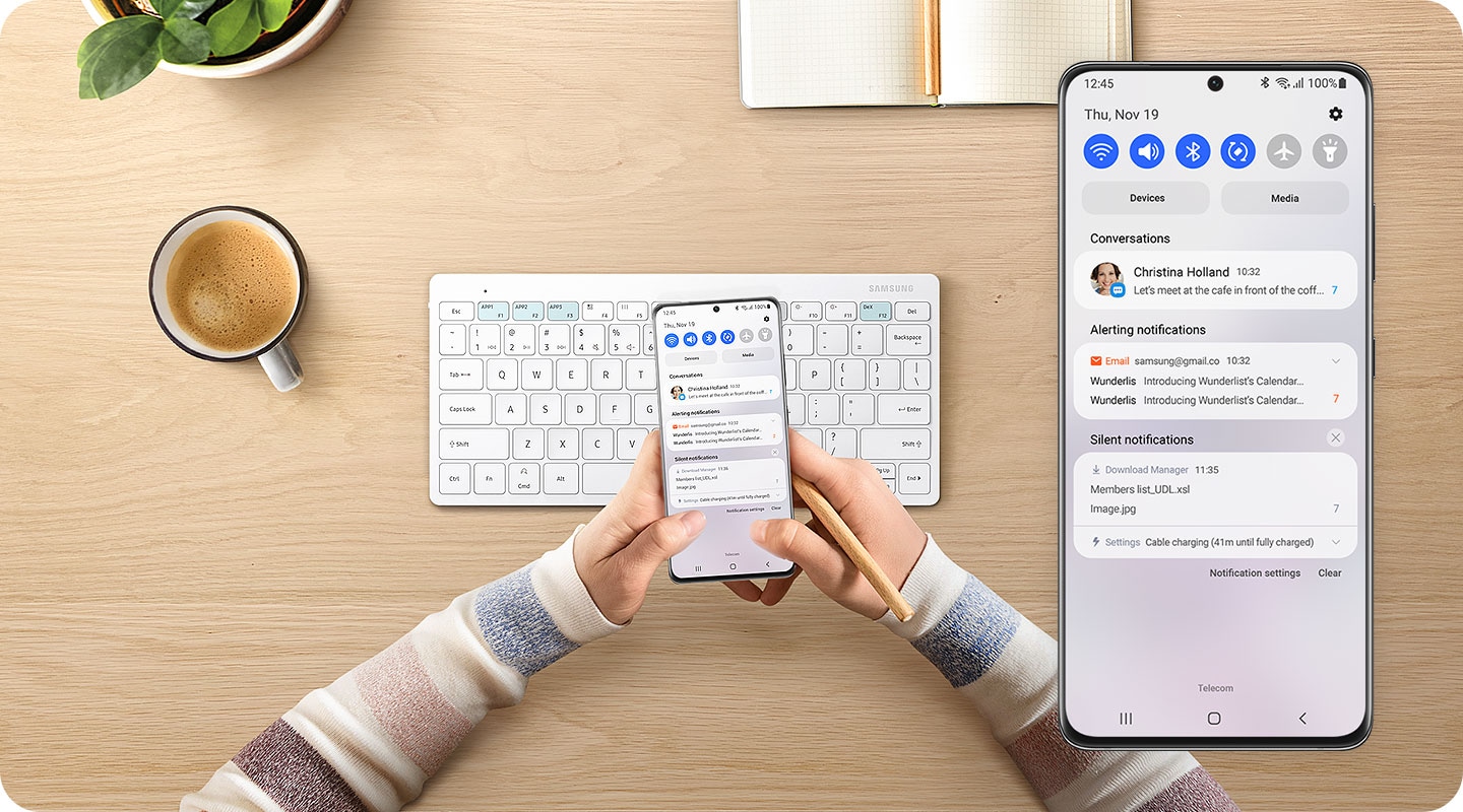 There is a white-colored Samsung Smart Keyboard Trio 500 on a desk. Above it, two hands are holding a smartphone seen from the front facing screen. On the right side, a close-up of on-screen of the phone is shown and the bluetooth mode indicator is on.