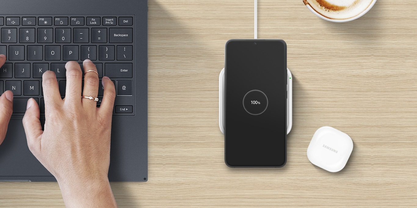 There is a closeup of a person’s hands typing on a laptop. Next to it, a black-colored Galaxy S22+ with the text 100% onscreen is on a power-connected 15W Wireless Charger Single. A white Galaxy buds2 case is placed next to the wireless charger on a wooden table.