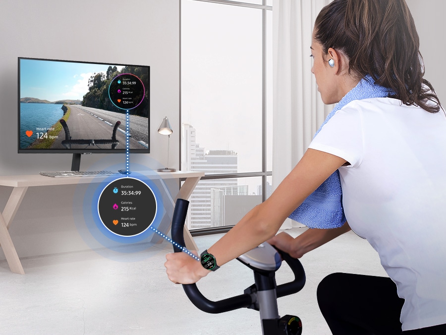 A woman is riding an indoor bicycle. She is wearing a Galaxy Watch and Buds, and her watch screen is overlaid on the monitor.