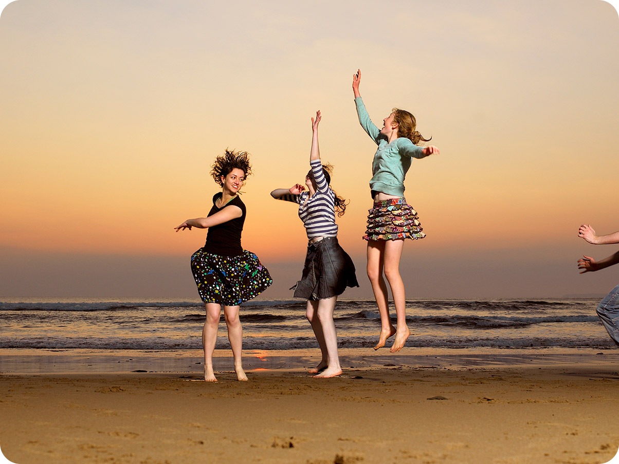1. Fotografija snimljena širokokutnom kamerom na kojoj se vide tri žene kako skaču na plaži pri zalasku sunca.