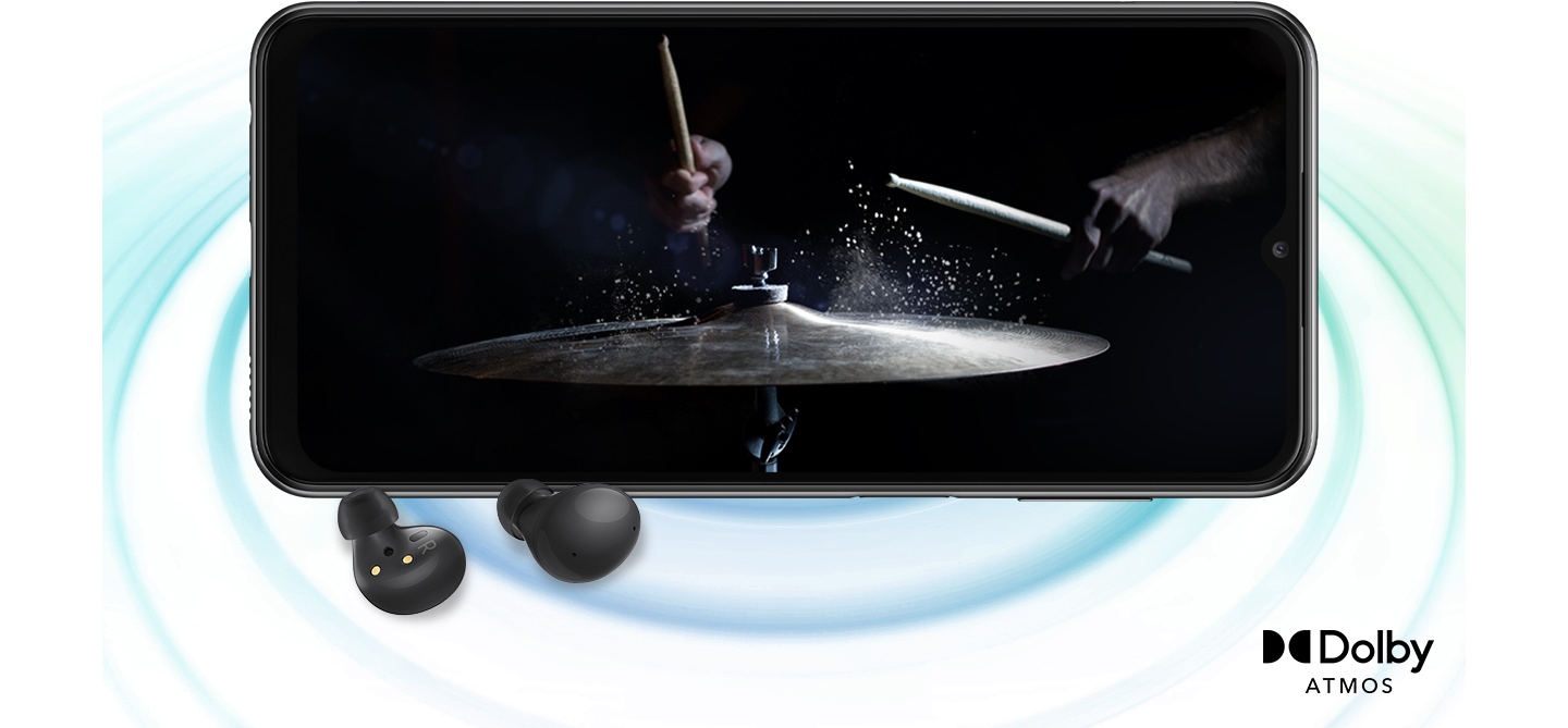 Galaxy A23 5G u pejzažnom načinu i slika s osobom koja svira bubnjeve u crnoj pozadini na zaslonu. Par crnih slušalica Galaxy Buds2 nalazi se ispred uređaja. Desno dolje je logo za Dolby Atmos.