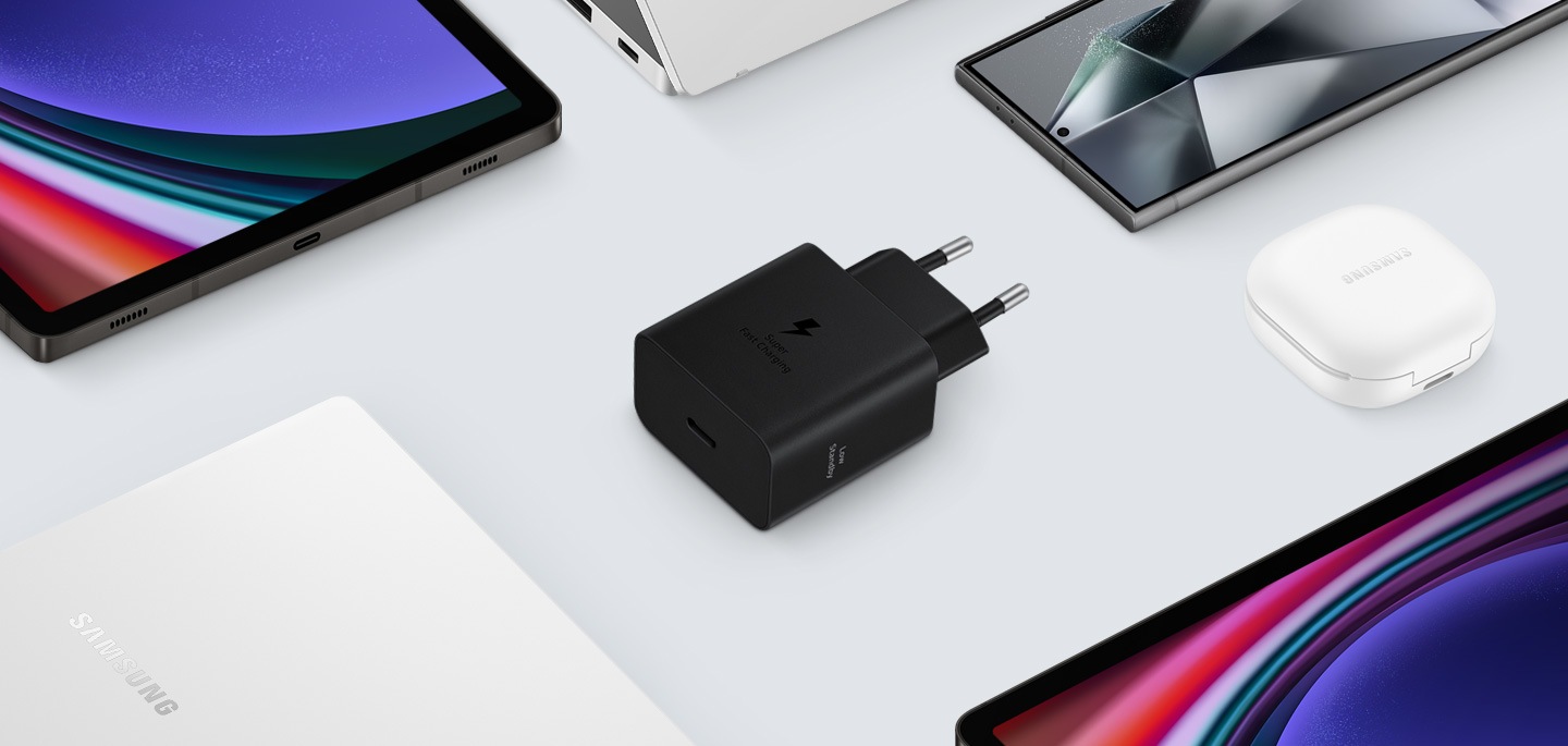 Prikazan je pogled odozgo na razne Samsung Galaxy uređaje s crnim adapterom za napajanje u sredini. Lijevo su prikazani Galaxy Book3, desno Galaxy Buds2 Pro u bijeloj boji, te Galaxy Tab S9 i Galaxy S24 Ultra.