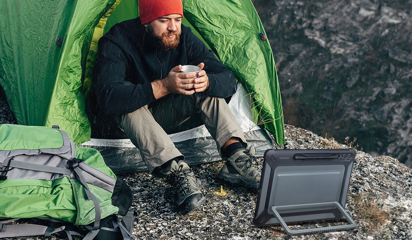 Egy bögrét tartó férfi ül egy kempingsátor előtt egy sziklás területen, és egy kültéri tokkal rendelkező Galaxy Tab S9+ készüléket néz, amely vízszintesen van elhelyezve a hátulján lévő állvánnyal.