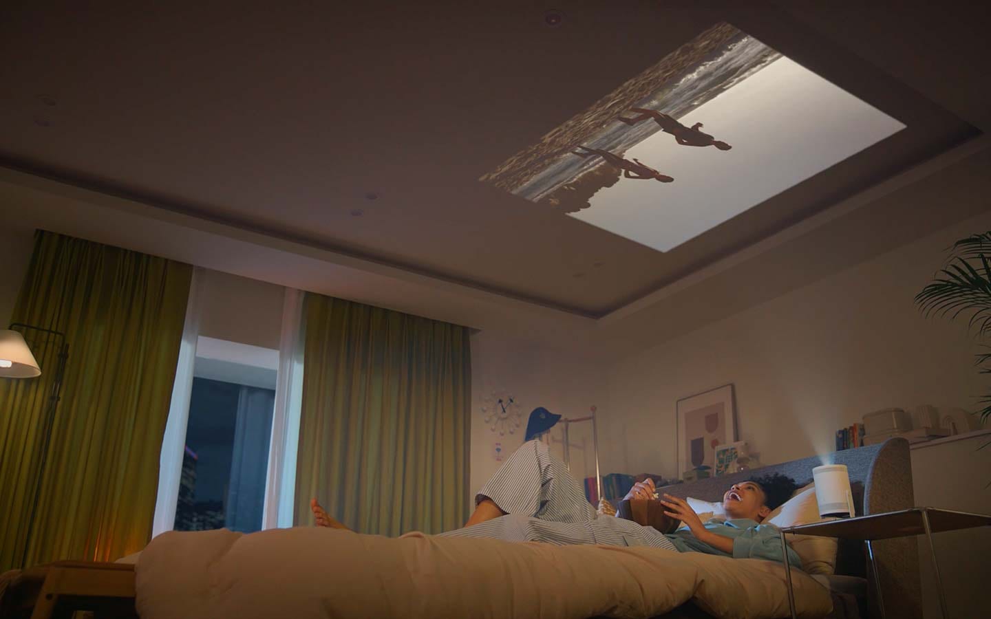 A woman is sitting on her bed eating popcorn, watching a video The Freestyle is projecting onto the wall in front of bed. Then she tilts The Freestyle so it points upwards and lays back with her head on the pillow, continuing to enjoying the video that’s now projected onto the ceiling.