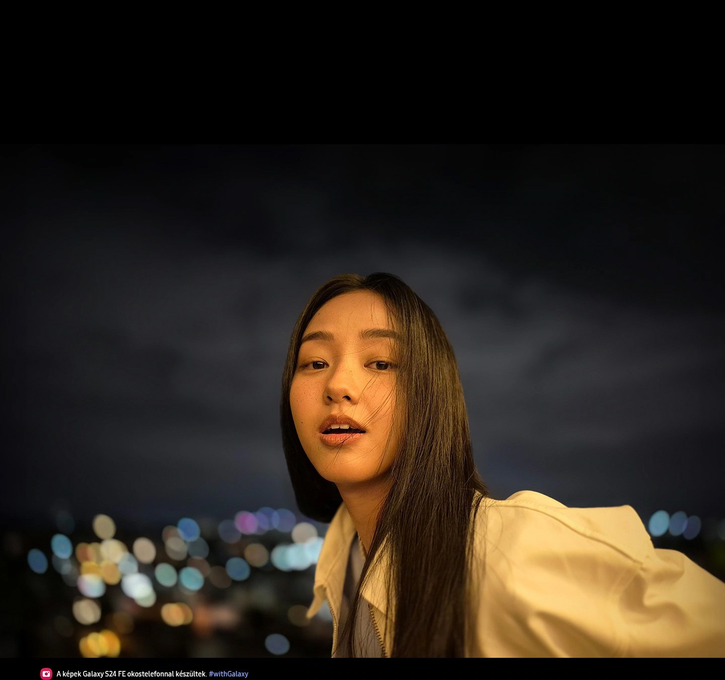 Egy nő fotója látható a szabadban éjszaka, kivilágított arccal és elmosódott városi fényekkel a háttérben. A szöveg a következő: A képek Galaxy S24 FE okostelefonnal készültek. #withGalaxy.