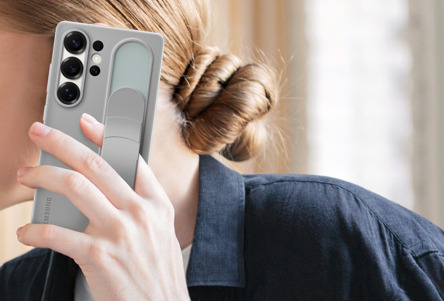 A woman holds a Galaxy S25 Ultra installed with the Standing Grip Case in Gray. One finger holding the phone is tucked under the adjustable grip.