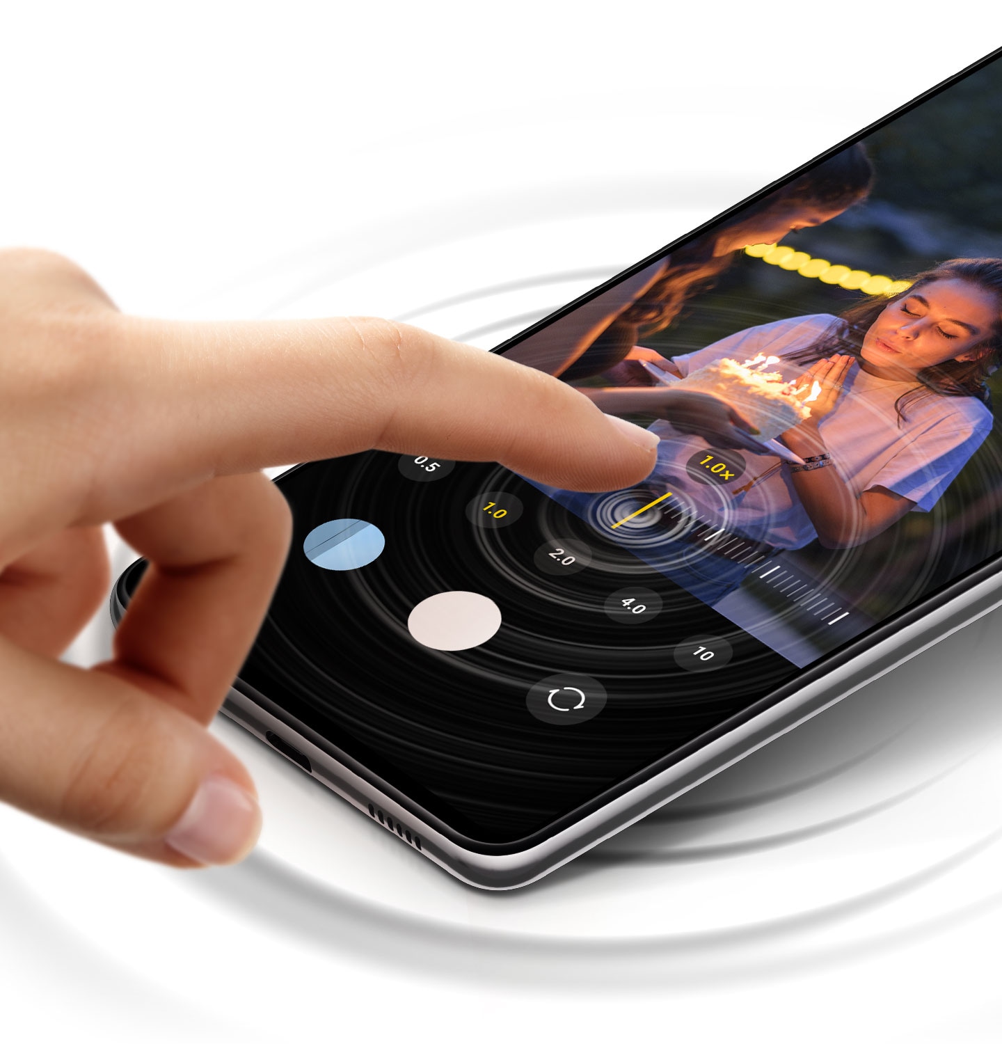 A Galaxy M54 5G shows a photo of a woman blowing out the candles on a cake. As a finger controls the camera zoom, haptic feedback is given, expressed as concentric circles over the phone.