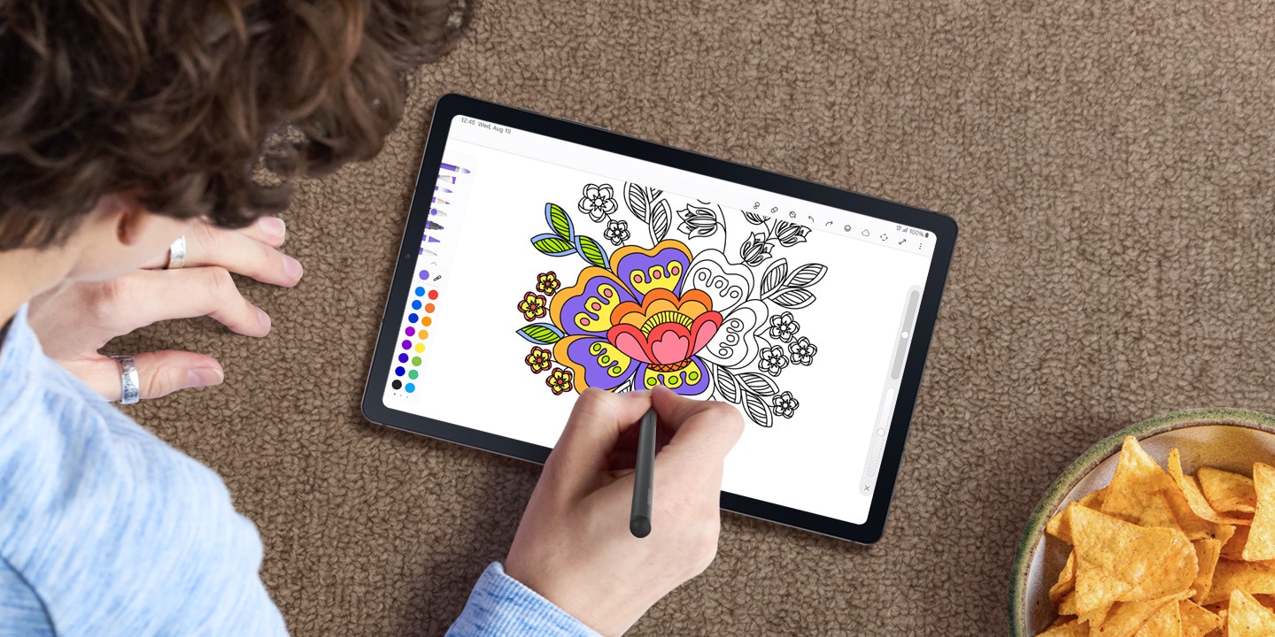Using the S Pen, a person is colouring an intricate image of a flower on the PENUP app on a Galaxy Tab S6 Lite device while sitting on a carpet floor with snacks on the side.