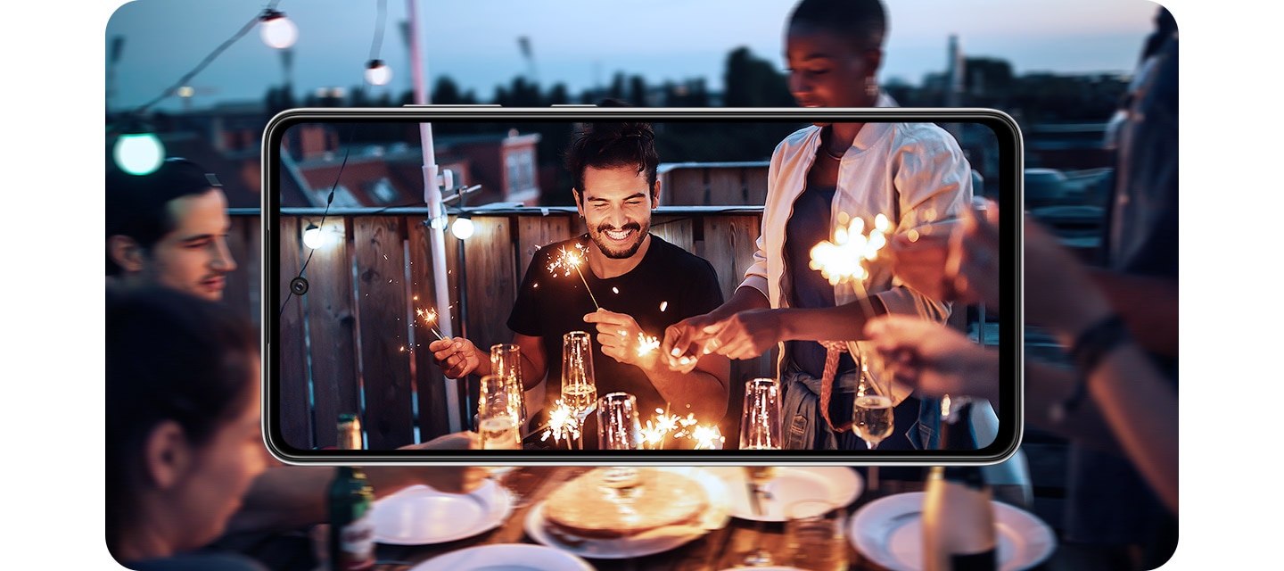 Galaxy A52 in landscape mode. Onscreen is a scene of people at a party in the evening playing with sparklers, and it expands outside of the display. The image inside of the display is brighter and more detailed than the portion outside of the display, demonstrating OIS.