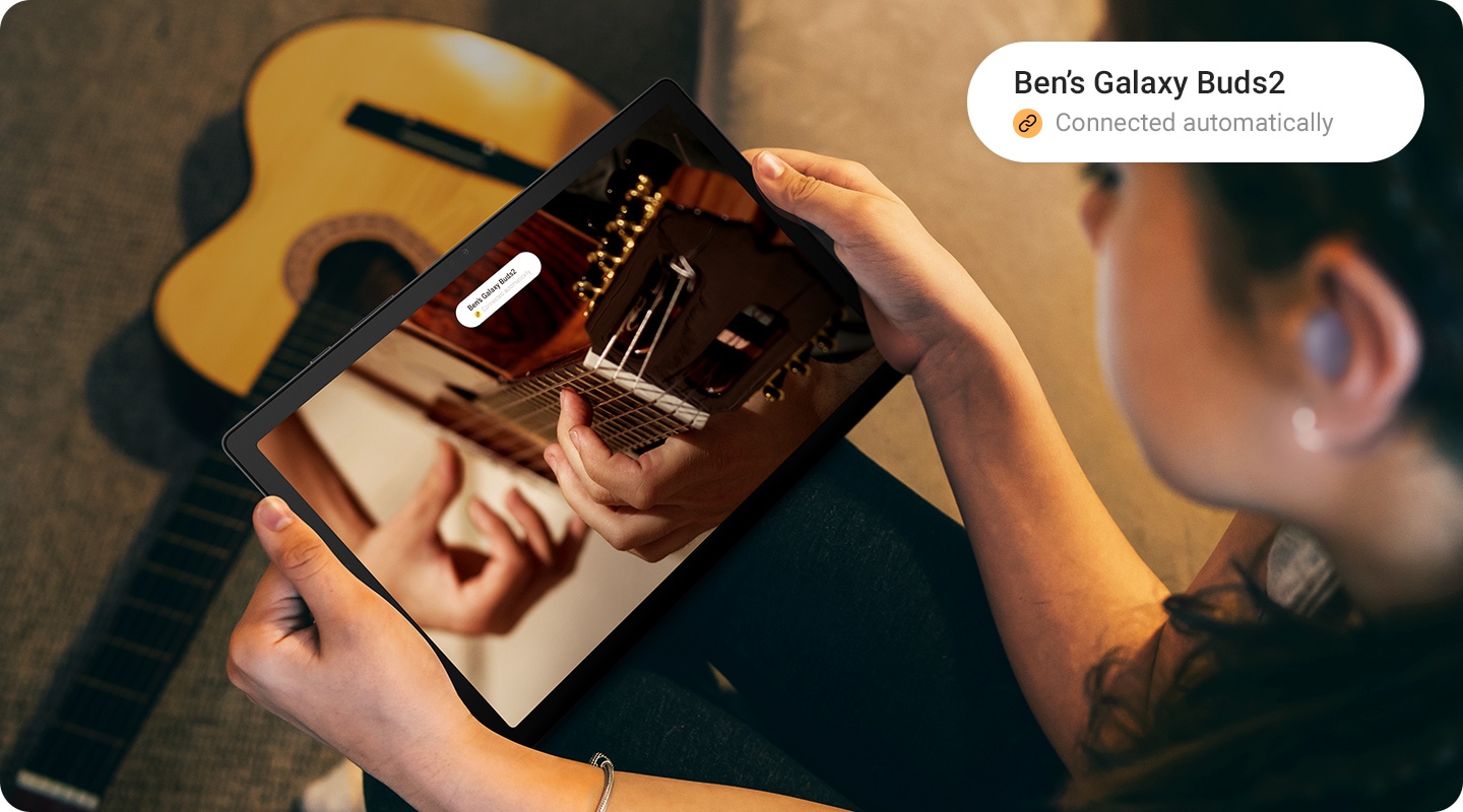 Woman wearing Galaxy Buds is watching a guitar playing video. Notification on Galaxy Tab A8 indicates the device owner's Galaxy Buds are automatically connected.