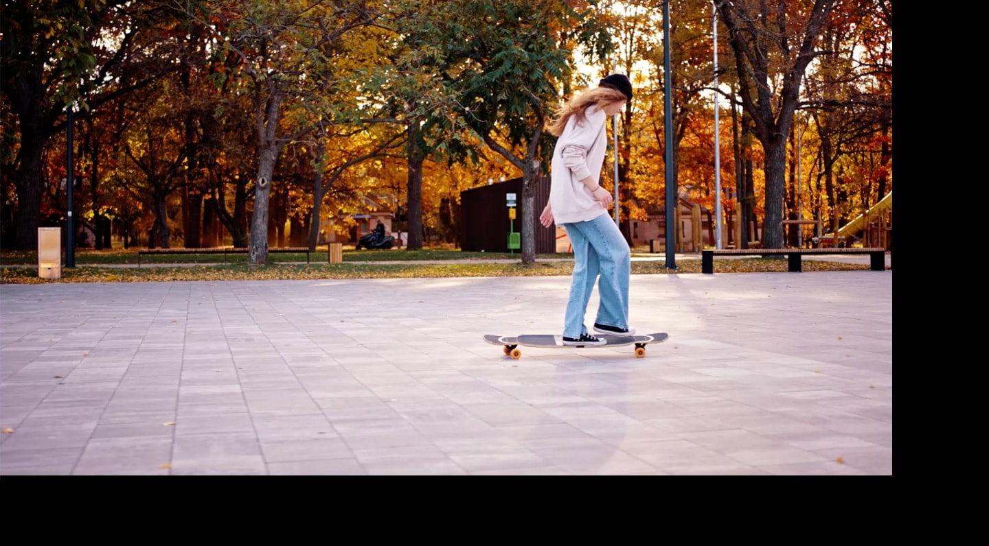 A long blonde haired skateboarder riding in a park by the forest.