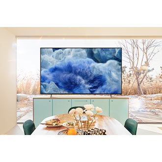 A QLED TV in a kitchen area displays a blue grassy landscape. Behind the TV, a glass window reveals the outdoor scenery.
