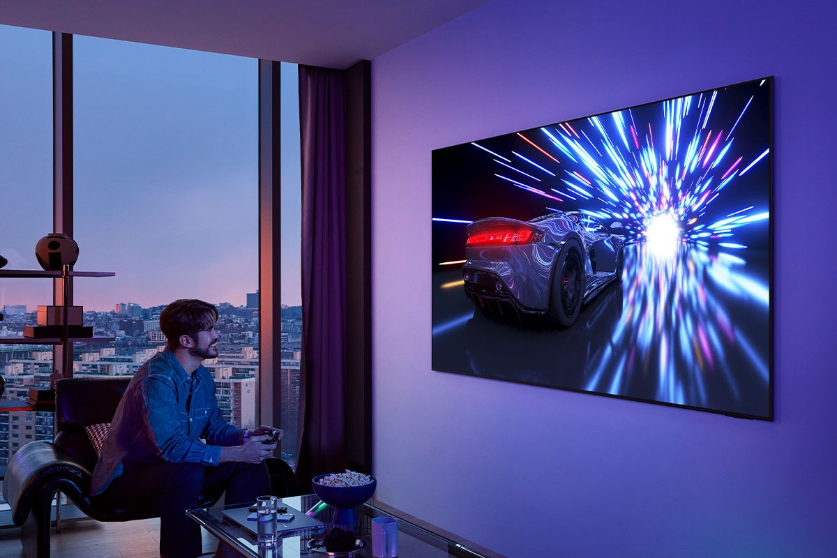 A man with a controller smiles as he games on a TV on the wall. The TV displays a racecar speeding through a lighted tunnel.