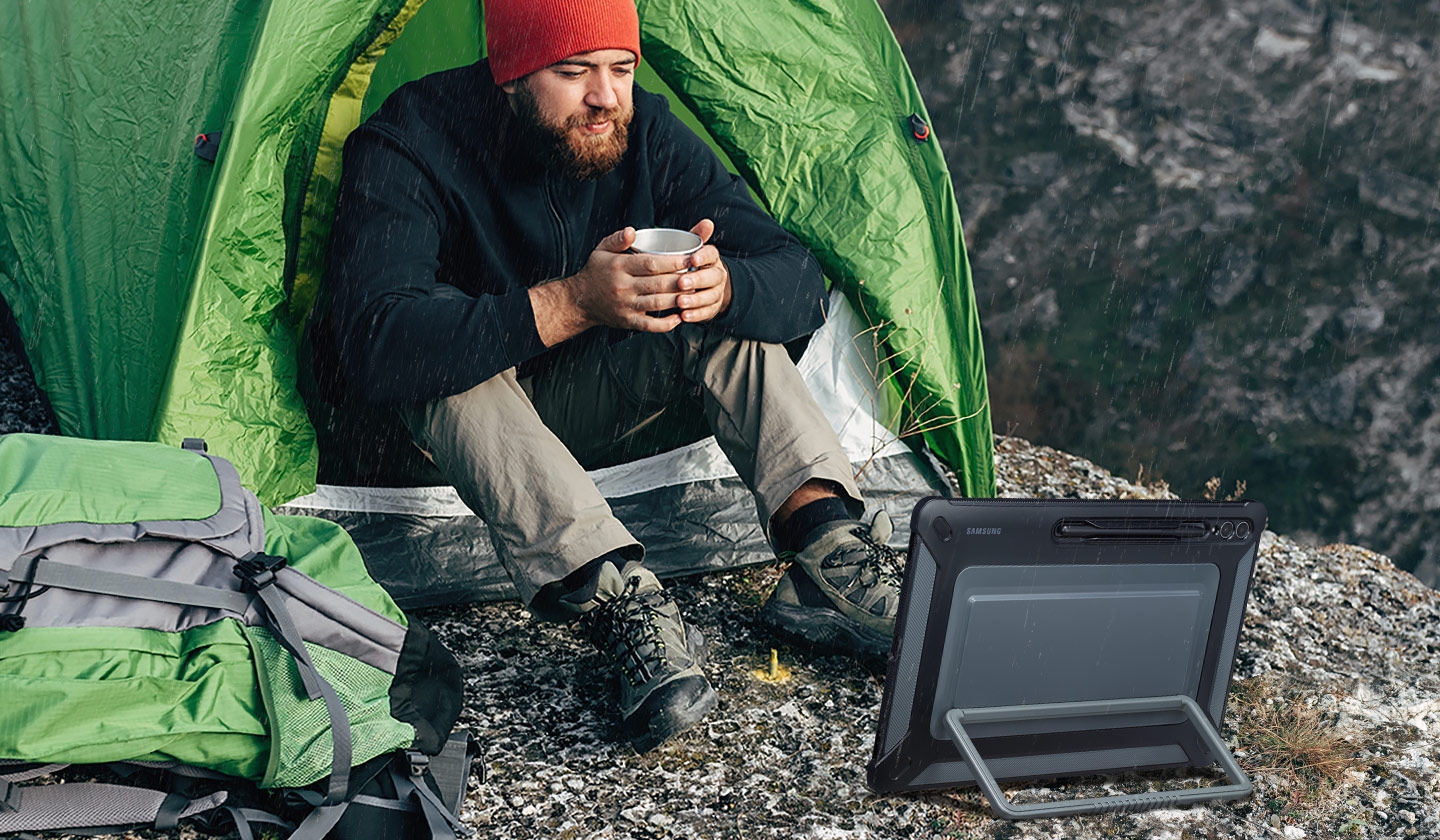 A man holding a mug is sitting in front of a camping tent on a rocky surface, looking at a Galaxy Tab S9 Ultra device with an Outdoor Cover on, placed horizontally using the kickstand on the back.