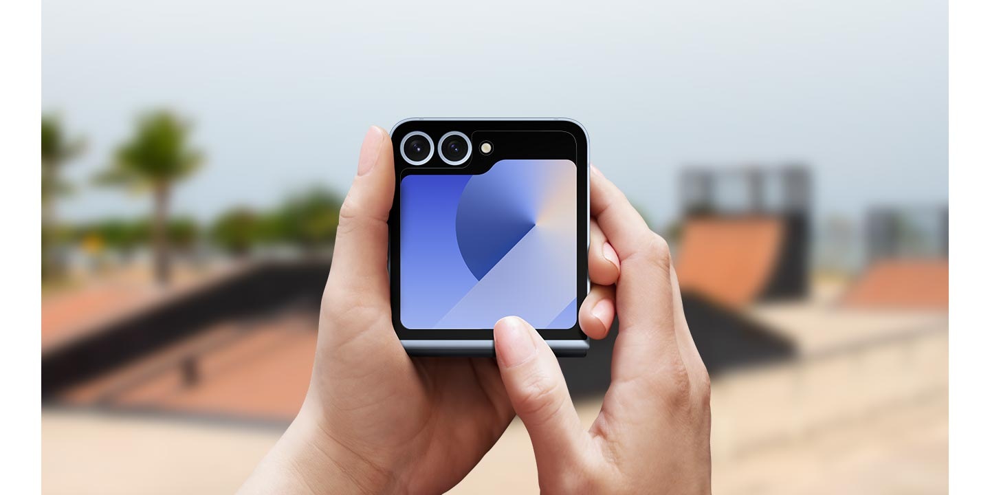 A person's hands holding the Galaxy Z Flip6 with an Anti-reflecting Film on the foreground. Their thumb is on the screen. The background is an out-of-focus skate park.