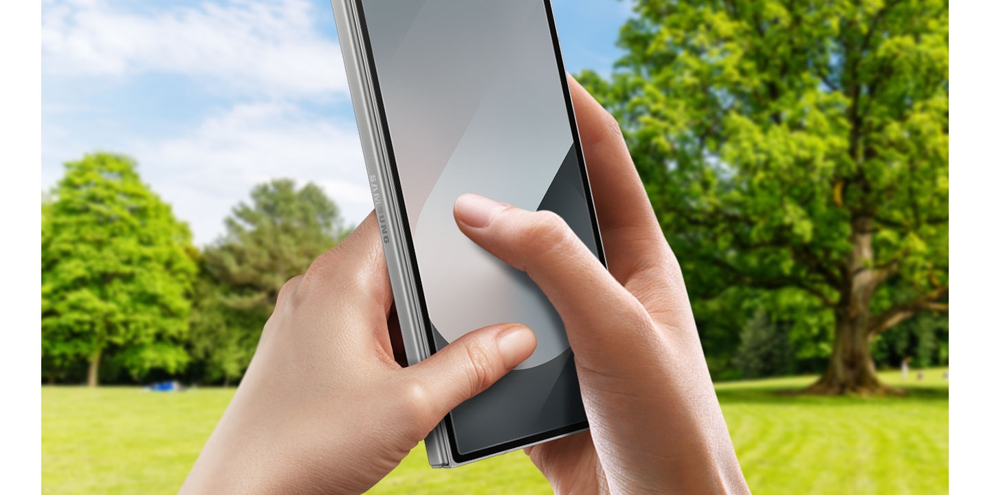 The background is a sunny park with green trees and fields. In the foreground, two hands hold a Galaxy Z Fold6. The thumbs are placed on the screen as if the phone was in use, showing off the high responsiveness of the display even with the Anti-reflecting Film.