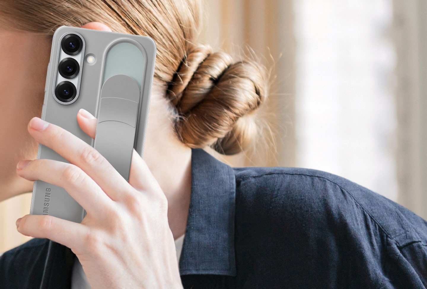 A woman holds a Galaxy S25 installed with the Standing Grip Case in Gray. One finger holding the phone is tucked under the adjustable grip.