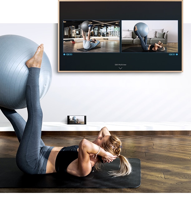 A woman is working out in front of her TV. On the TV screen she can see a screen of herself as well as the screen of the fitness trainer and below the TV her smartphone camera is capturing her workout so she can see it on the TV scree .