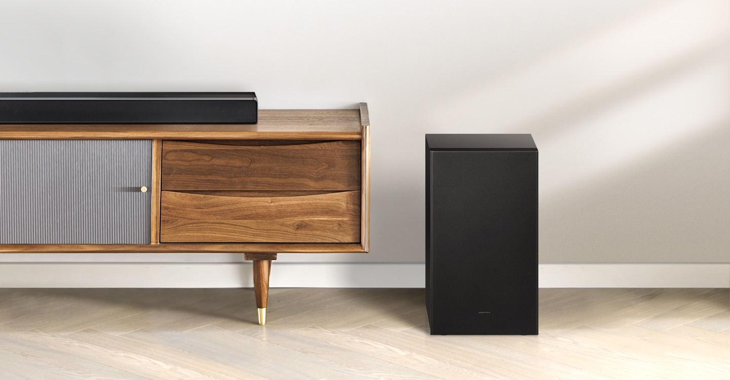 Samsung A series soundbar is being shown on top of a contemporary TV cabinet along with its matching subwoofer which is being displayed to the side of the TV cabinet.