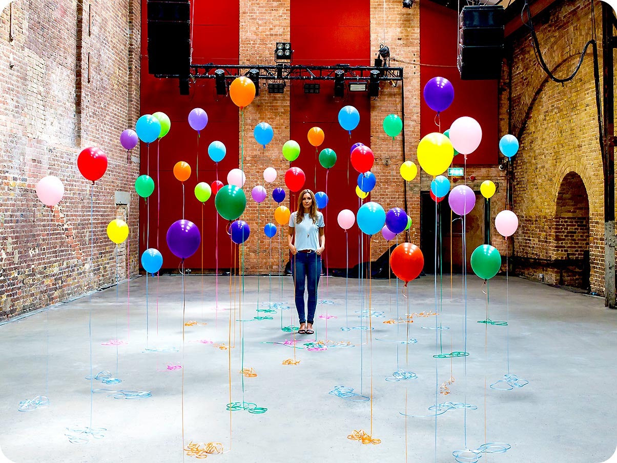 2. There is a woman standing in the middle of many different color balloons. With Ultra Wide Camera, you can see all of the balloons surrounding her as well as more of the stage background.