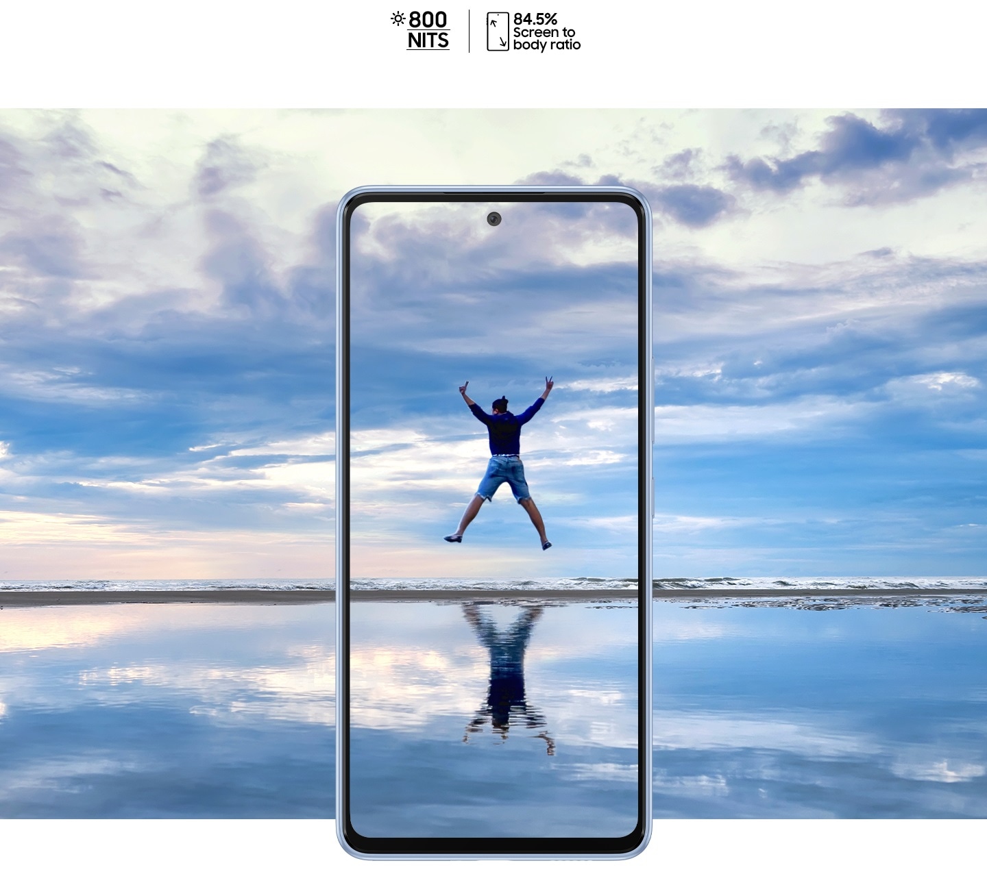 galaxy a53 5g seen from the front against a beautiful landscape that overlaps onto the screen. it shows an expansive sky over water that reflects it with a thin horizon dissecting near the middle. in the center of the screen, a man is jumping in the air with all four limps outstretched and his reflection is shown on the water.