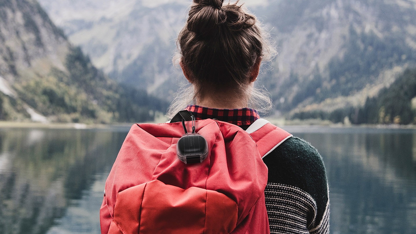 A person outside overlooking a lake. Galaxy Buds Pro are inside the Protective cover and clipped onto the backpack's handle with the included carabiner.