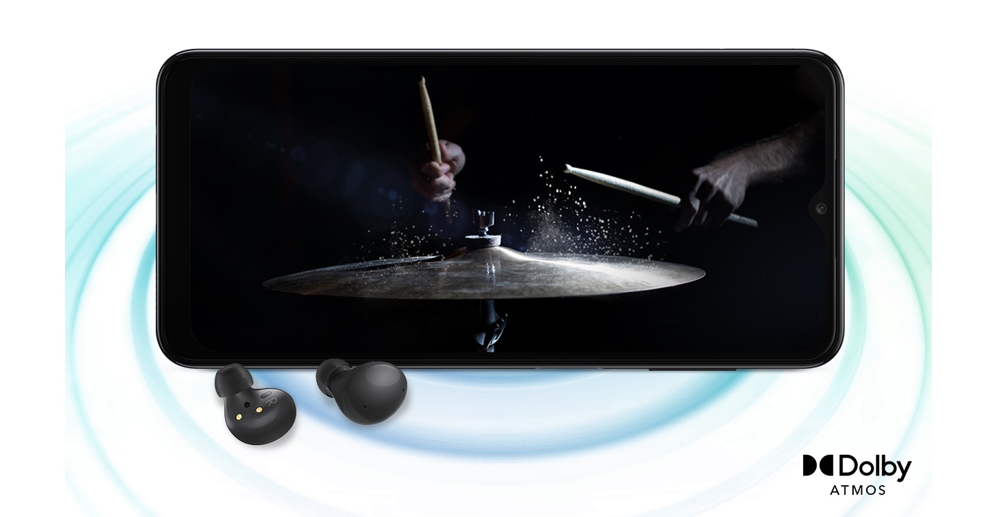 Galaxy A04s in landscape mode and an image with a person playing drums in the black background onscreen. A pair of black Galaxy Buds2 are placed in front of the device. On the right bottom is a logo for Dolby Atmos.