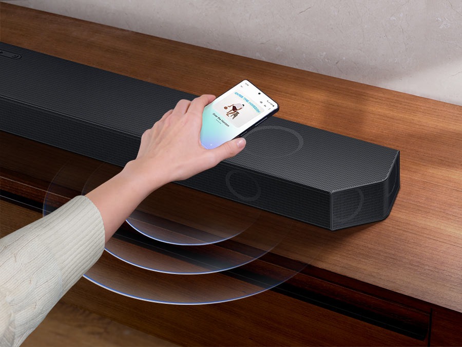 A hand taps a smartphone with the Samsung music app on-screen on the Soundbar. It instantly plays music, showing how easy it is to switch from smartphone to Soundbar.