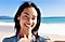 A selfie of a female smiling, holding the device with left hand. Behind her is a bright, blurry view of the beach and blue sky.
