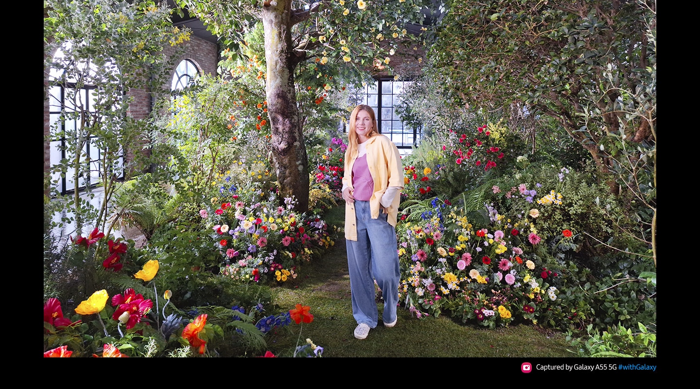 A photo taken with 50 Megapixels of high-resolution of a person standing in a lush indoor garden full of colorful flowers and greenery. Text reads Captured by Galaxy A55 5G #withGalaxy.