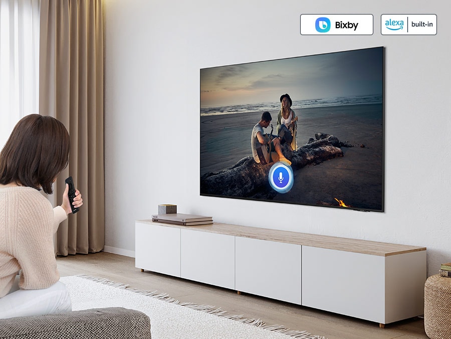 A woman uses her voice assistant to control her TV. "Bixby" and "Alexa built-in" logos.
