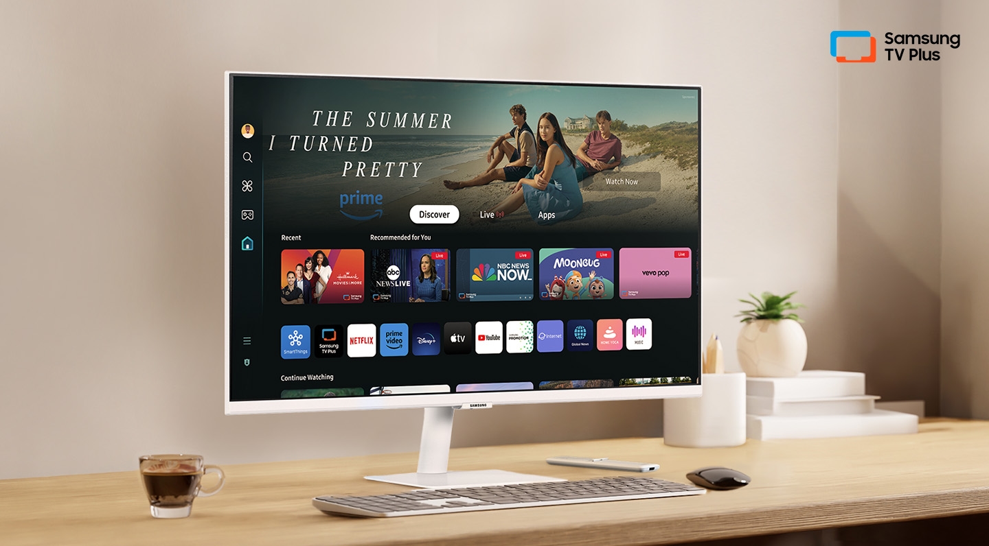 There is a monitor on a desk and its home UI is shown with OTT applications. On the upper right, there's a logo of Samsung TV Plus.