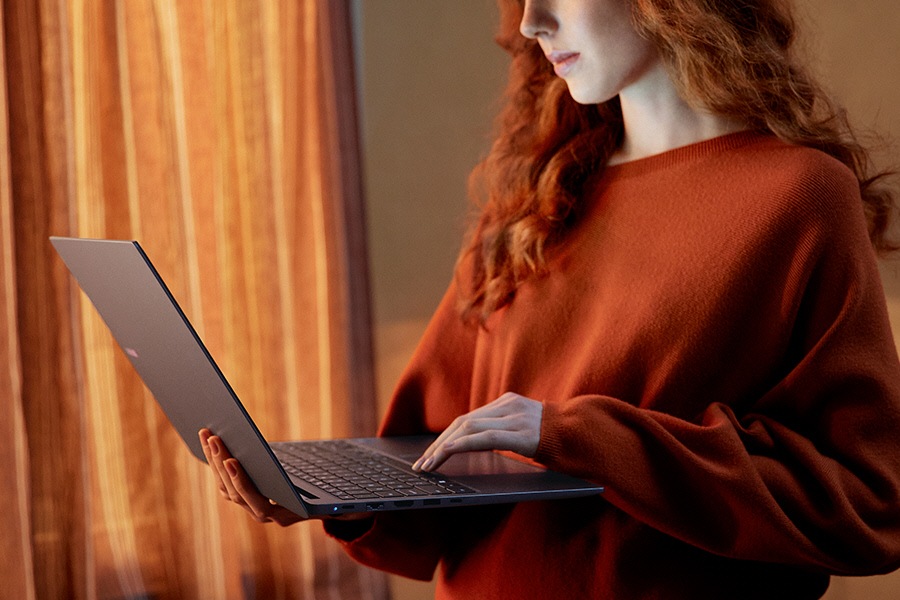 A young woman standing indoors is holding Galaxy Book4 in Gray open while typing on the laptop keyboard.