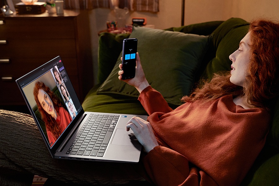 A young woman sitting on a sofa at home is using Galaxy S24 Plus as a connected camera with Galaxy Book4 in Gray to participate in a Microsoft Teams group video call with three other people shown onscreen.